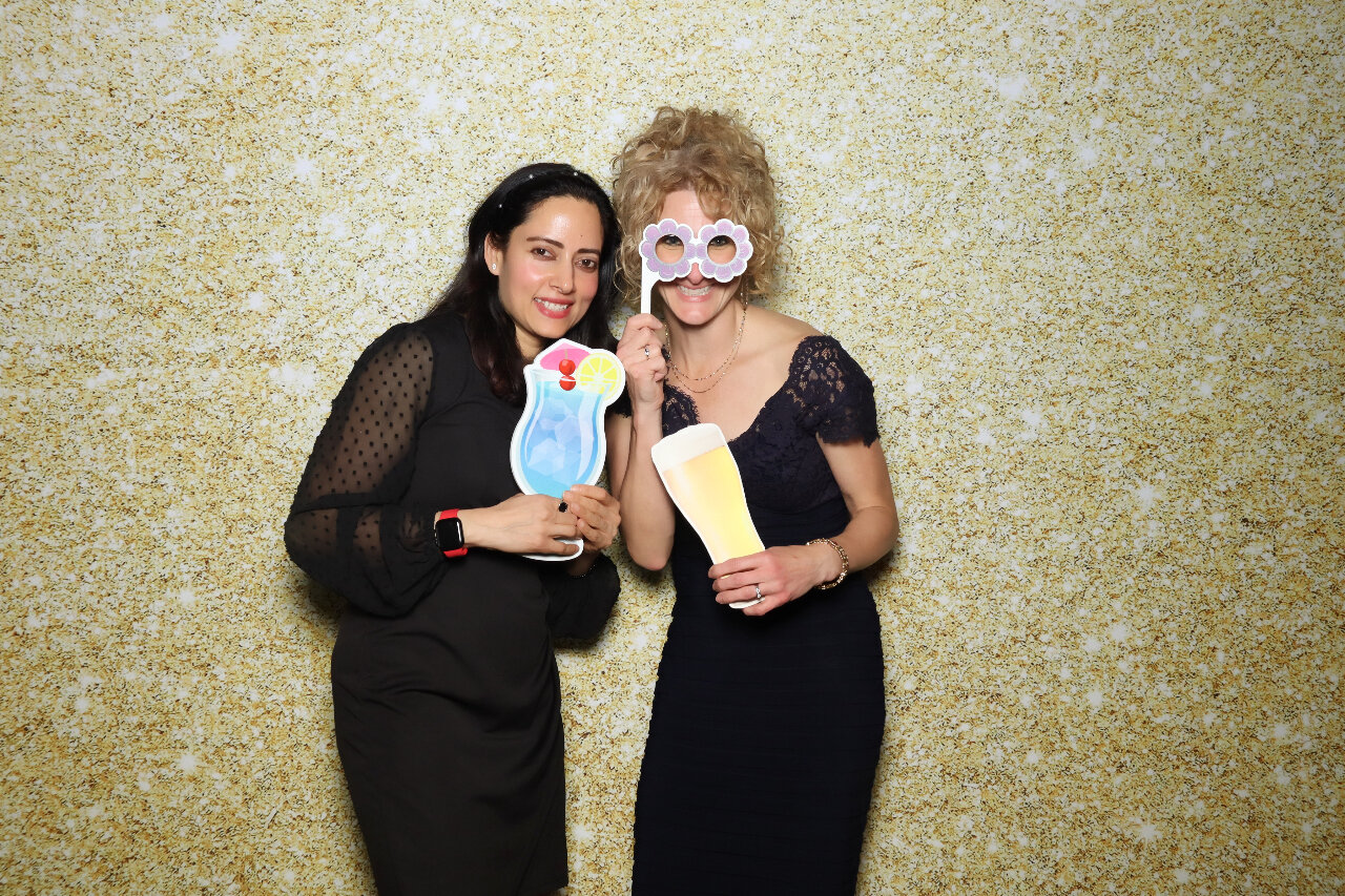 two women posing together and holding props - one is holding a colorful drink and the other is holding a large flower-shaped prop. They are both smiling and dressed in black outfits. The background is a shimmery gold/